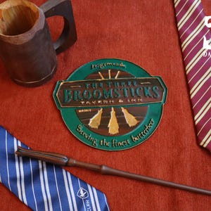 K&ouml;nnte beinhalten: Eine Auswahl an thematischen Artikeln, darunter ein Holzkrug, ein Zauberstab und zwei gestreifte Krawatten. Ein rundes Schild zeigt "The Three Broomsticks Tavern & Inn". Die Krawatten sind blau und bordeaux mit goldenen Streifen. Auf roter Oberfl&auml;che.