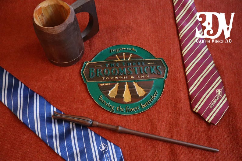 K&ouml;nnte beinhalten: Eine Auswahl an thematischen Artikeln, darunter ein Holzkrug, ein Zauberstab und zwei gestreifte Krawatten. Ein rundes Schild zeigt "The Three Broomsticks Tavern & Inn". Die Krawatten sind blau und bordeaux mit goldenen Streifen. Auf roter Oberfl&auml;che.