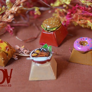 Puede incluir: Cuatro teclas de miniatura de comida, cada una con un diseño de comida diferente: una rebanada de pizza, un tazón de fideos, una hamburguesa y una dona rosada con chispas de colores. Las teclas están sobre una superficie marrón.