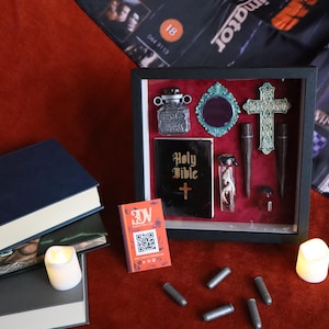 May include: A framed display of gothic and religious items, including a small Bible, a cross, and a mirror. Other decorative objects are also present. Several books and a small orange card with a QR code are also visible. The items are arranged on a red surface.