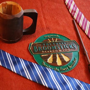 K&ouml;nnte beinhalten: Ein Holzbecher, zwei gestreifte Krawatten, ein Zauberstab und ein Schild f&uuml;r The Three Broomsticks Tavern & Inn werden auf einer roten Oberfl&auml;che pr&auml;sentiert. Die Krawatten sind blau und wei&szlig; sowie bordeauxrot und goldfarben. Das Schild ist gr&uuml;n und braun.