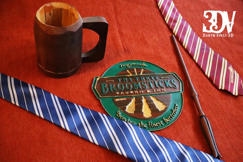 K&ouml;nnte beinhalten: Ein Holzbecher, zwei gestreifte Krawatten, ein Zauberstab und ein Schild f&uuml;r The Three Broomsticks Tavern & Inn werden auf einer roten Oberfl&auml;che pr&auml;sentiert. Die Krawatten sind blau und wei&szlig; sowie bordeauxrot und goldfarben. Das Schild ist gr&uuml;n und braun.