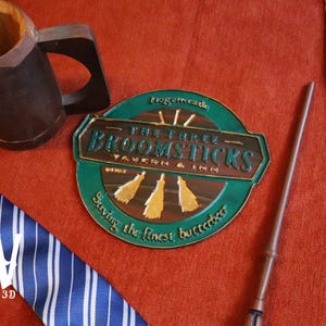 K&ouml;nnte beinhalten: Eine Auswahl an thematischen Gegenst&auml;nden auf roter Oberfl&auml;che. Enth&auml;lt einen Holzkrug, einen Zauberstab, ein Schild f&uuml;r "The Three Broomsticks Tavern & Inn" und zwei gestreifte Krawatten, eine blaue und eine weinrote.