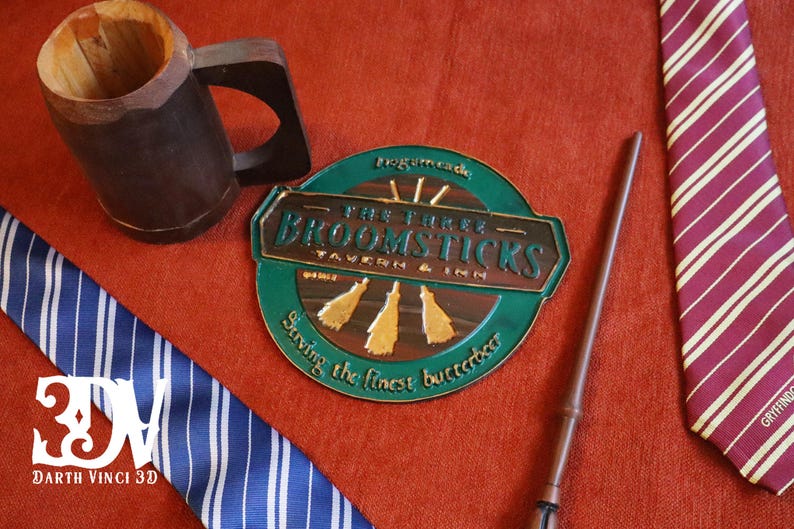 K&ouml;nnte beinhalten: Eine Auswahl an thematischen Gegenst&auml;nden auf roter Oberfl&auml;che. Enth&auml;lt einen Holzkrug, einen Zauberstab, ein Schild f&uuml;r "The Three Broomsticks Tavern & Inn" und zwei gestreifte Krawatten, eine blaue und eine weinrote.