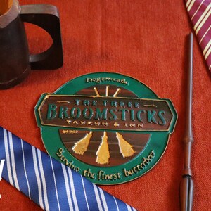 K&ouml;nnte beinhalten: Eine Auswahl an thematischen Artikeln, darunter ein Holzkrug, ein Zauberstab und zwei gestreifte Krawatten in Blau und Burgunderrot. Ein rundes Schild f&uuml;r die Taverne "The Three Broomsticks" ist ebenfalls vorhanden, mit dem Text "Serving the finest butterbeer."