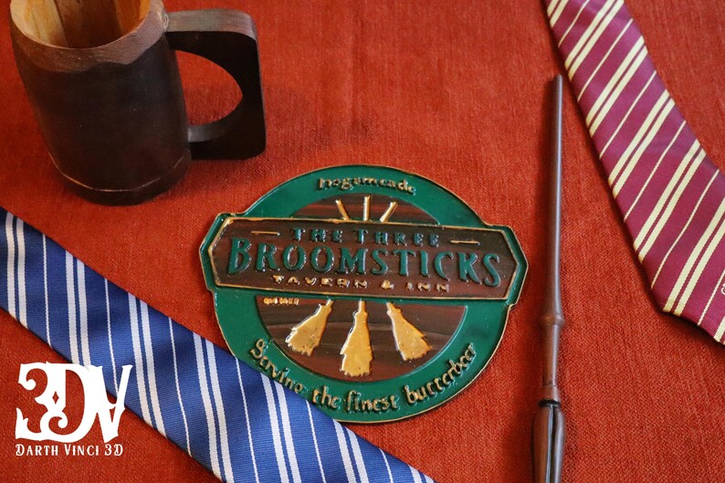 K&ouml;nnte beinhalten: Eine Auswahl an thematischen Artikeln, darunter ein Holzkrug, ein Zauberstab und zwei gestreifte Krawatten in Blau und Burgunderrot. Ein rundes Schild f&uuml;r die Taverne "The Three Broomsticks" ist ebenfalls vorhanden, mit dem Text "Serving the finest butterbeer."