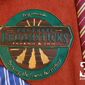 K&ouml;nnte beinhalten: Ein kreisf&ouml;rmiges, gr&uuml;n-braunes Schild f&uuml;r "The Three Broomsticks Tavern & Inn" mit goldenen Akzenten und Text. Das Schild befindet sich auf einem roten, strukturierten Stoff, mit einer gestreiften Krawatte im Hintergrund.