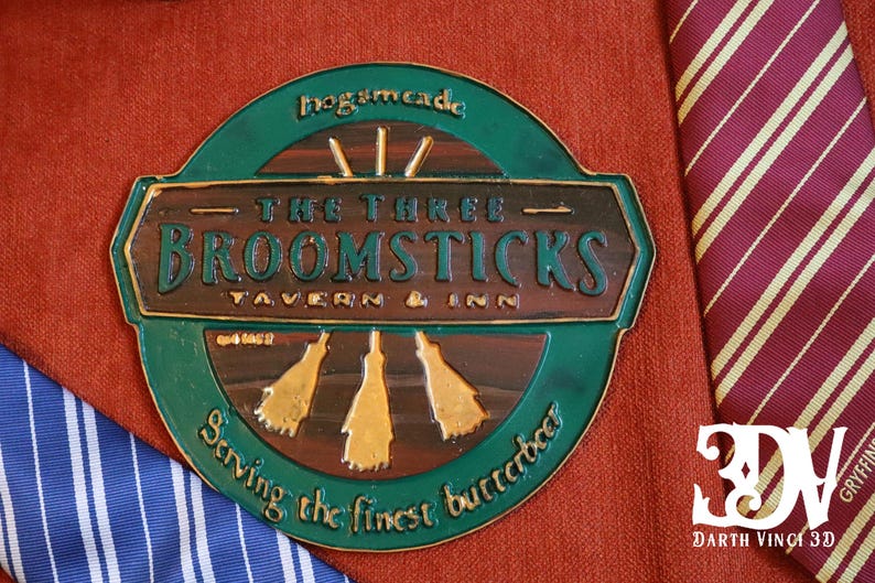 K&ouml;nnte beinhalten: Ein kreisf&ouml;rmiges, gr&uuml;n-braunes Schild f&uuml;r "The Three Broomsticks Tavern & Inn" mit goldenen Akzenten und Text. Das Schild befindet sich auf einem roten, strukturierten Stoff, mit einer gestreiften Krawatte im Hintergrund.