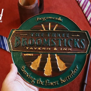 K&ouml;nnte beinhalten: Ein gr&uuml;nes und braunes Schild f&uuml;r "The Three Broomsticks Tavern & Inn" mit goldenen Akzenten. Das Schild ist rund mit den Worten "Hogsmeade" oben und "Serving the finest butterbeer" unten. Ein Zauberstab und Krawatten sind im Hintergrund.
