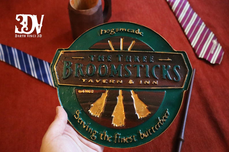 K&ouml;nnte beinhalten: Ein gr&uuml;nes und braunes Schild f&uuml;r "The Three Broomsticks Tavern & Inn" mit goldenen Akzenten. Das Schild ist rund mit den Worten "Hogsmeade" oben und "Serving the finest butterbeer" unten. Ein Zauberstab und Krawatten sind im Hintergrund.