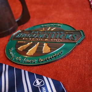 K&ouml;nnte beinhalten: Ein ovales Schild mit dem Text "Broomsticks Tavern & Inn" in metallischen Buchstaben, mit einem gr&uuml;nen und braunen Farbschema. Das Schild befindet sich auf einer roten Oberfl&auml;che, mit einem braunen Krug und gestreiften Krawatten im Hintergrund.
