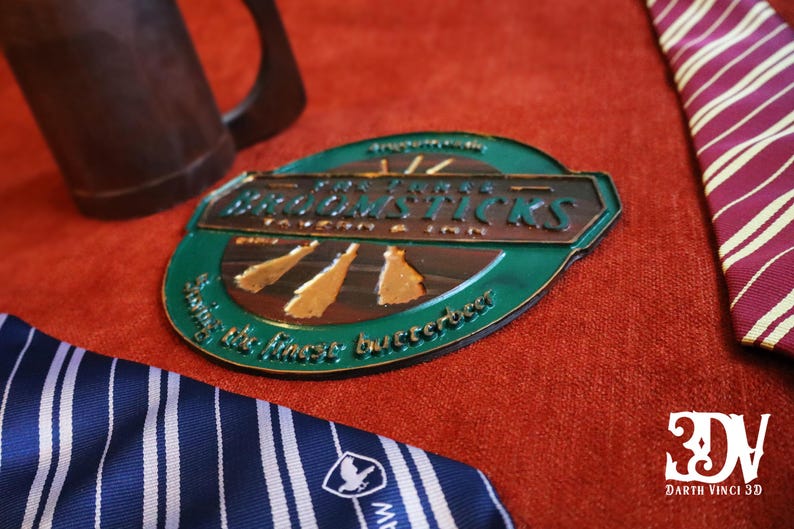 K&ouml;nnte beinhalten: Ein ovales Schild mit dem Text "Broomsticks Tavern & Inn" in metallischen Buchstaben, mit einem gr&uuml;nen und braunen Farbschema. Das Schild befindet sich auf einer roten Oberfl&auml;che, mit einem braunen Krug und gestreiften Krawatten im Hintergrund.
