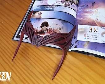 May include: A dark red, pointed headpiece with a symmetrical, wing-like design and recessed detailing. The accessory features a sharp, angular silhouette with two long, downward-curving side points and a central peak, crafted in a matte finish.