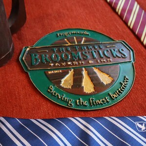 K&ouml;nnte beinhalten: Ein gr&uuml;nes und bronzefarbenes Metallschild f&uuml;r "The Three Broomsticks Tavern & Inn" mit dem Text "Serving the finest butterbeer". Ein brauner Krug und gestreifte Krawatten sind ebenfalls sichtbar.