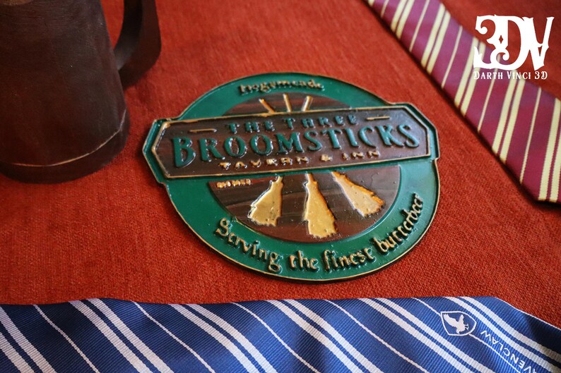 K&ouml;nnte beinhalten: Ein gr&uuml;nes und bronzefarbenes Metallschild f&uuml;r "The Three Broomsticks Tavern & Inn" mit dem Text "Serving the finest butterbeer". Ein brauner Krug und gestreifte Krawatten sind ebenfalls sichtbar.