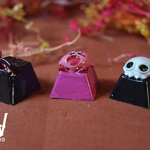 Puede incluir: Tres teclas personalizadas pintadas con un personaje rosa de Kirby, una lengua roja y negra y una calavera blanca con ojos verdes. Las teclas están sobre bases negras y rosas.