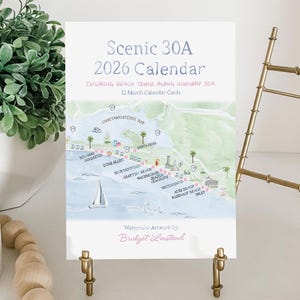 May include: A 2026 calendar titled "Scenic 30A" featuring a watercolor map of beach towns along Highway 30A. The calendar is displayed on a gold easel, with a green plant and wooden beads in the background.