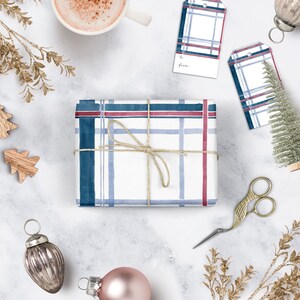 Pu&ograve; includere: Una scatola regalo bianca avvolta in carta a quadri blu e rossi con un fiocco di spago marrone. La scatola &egrave; circondata da varie decorazioni natalizie, tra cui un piccolo albero di Natale, ornamenti e una tazza di caff&egrave;.