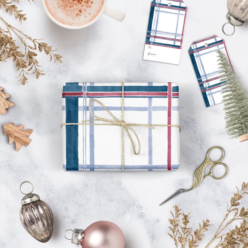 Pu&ograve; includere: Una scatola regalo bianca avvolta in carta a quadri blu e rossi con un fiocco di spago marrone. La scatola &egrave; circondata da varie decorazioni natalizie, tra cui un piccolo albero di Natale, ornamenti e una tazza di caff&egrave;.