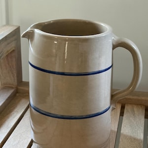 May include: A cylindrical ceramic pitcher with a glossy beige finish and two horizontal dark blue stripes encircling the body. The pitcher features a curved handle on the right side and a small pouring spout at the top rim, showcasing a smooth texture.