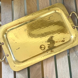 May include: A rectangular brass serving tray with a hammered finish and two twisted rope-style handles attached with copper rivets. The surface of the tray shows signs of wear and patina, with visible dark spots and a reflective metallic gold tone.
