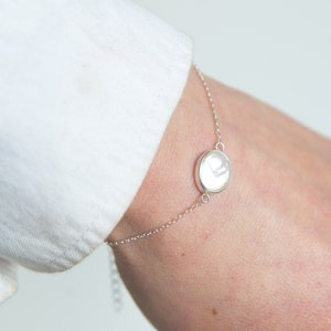 May include: A delicate silver bracelet with a round, white pendant. The bracelet is worn on a wrist, with a white shirt sleeve in the background. The jewellery is simple and elegant, suitable for everyday wear. The pendant is approximately 1 cm in diameter.