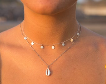 May include: A layered silver necklace with a shell pendant and small disc charms. The necklace is worn against a person's skin, with a yellow top visible in the background. The necklace is a delicate piece of jewelry.