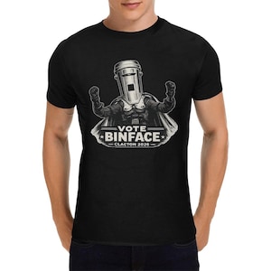 Op de afbeelding: Een zwart T-shirt met korte mouwen en een afbeelding van een personage met een prullenbak als hoofd, een cape dragend en beide vuisten omhoog. Onder het personage staat de tekst VOTE BINFACE CLACTON 2026 in witte hoofdletters.