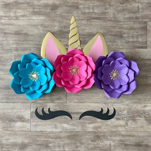 May include: A unicorn head made of paper flowers. The unicorn has a pink nose, a gold horn, and blue and purple flowers for ears. The unicorn has black eyelashes.