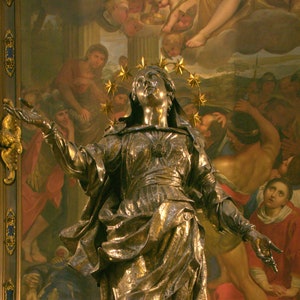 May include: A gold-coloured statue of the Virgin Mary with a crown of stars, standing on a crescent moon. The statue is surrounded by a painting depicting a scene from the Bible.