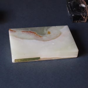 Puede incluir: Caja rectangular de ónix con un patrón veteado en verde claro y blanco, acentuado con vetas marrones y doradas. La caja tiene un acabado natural y pulido y una pequeña etiqueta rectangular con texto.