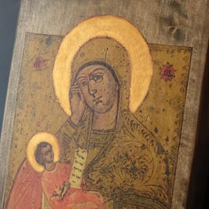 May include: A vintage religious icon featuring a depiction of a woman with a golden halo, holding a child. The artwork is painted on a wooden panel with a distressed, aged appearance, showcasing a traditional style with muted colors.