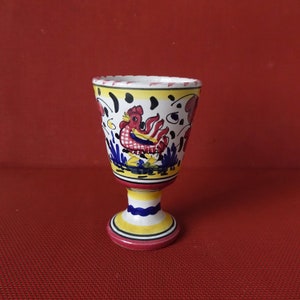 May include: A white ceramic goblet with a yellow, blue and red rooster design. The goblet has a wide base and a narrow top.