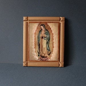 May include: A framed image of Our Lady of Guadalupe, a religious icon, in a gold frame. The image depicts the Virgin Mary in a blue and gold robe, standing on a crescent moon.