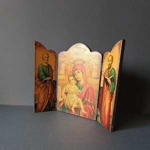 May include: A wooden triptych featuring religious icons. The central panel depicts the Virgin Mary and Jesus, flanked by images of saints. The artwork has a gold background and is framed in a distressed, antique style.