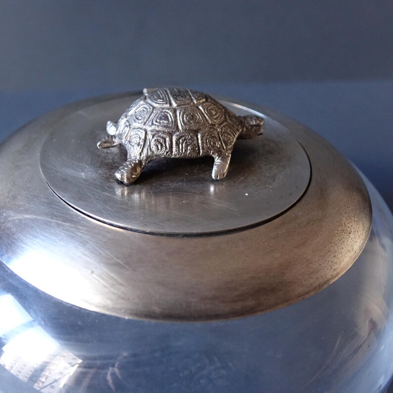 May include: A silver lidded jar with a silver turtle figurine on top. The jar is made of clear glass and has a silver lid.
