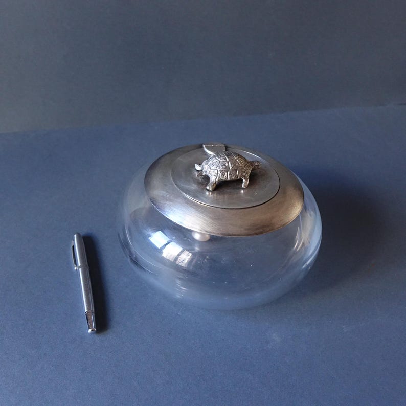 May include: A clear glass jar with a silver lid featuring a turtle figurine. The jar is round and has a smooth surface.