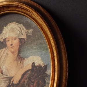 Può includere: Un ritratto ovale incorniciato di una donna con un berretto bianco e un vestito bianco. Sta tenendo un cavallo marrone con una briglia bianca. La cornice è dorata.