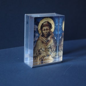 Puede incluir: Un cubo de vidrio transparente con una imagen impresa de un hombre con una túnica marrón y un halo. El fondo es azul y dorado.