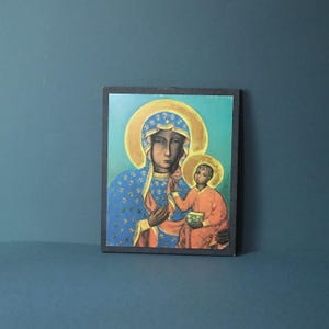 Pode incluir: Uma placa de parede retangular com uma pintura de ícone religioso de uma figura em uma capa azul com padrões de flor-de-lis dourados e um halo dourado, segurando uma criança em uma vestimenta vermelha com um halo dourado correspondente em um fundo azul-petróleo.