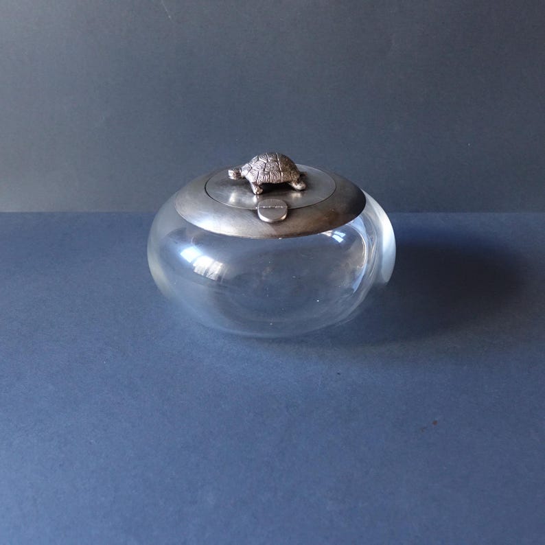 May include: A clear glass jar with a silver lid featuring a small turtle figurine. The jar is round and has a smooth surface.