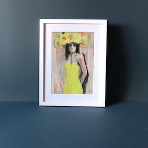 May include: Framed artwork depicting a woman in a yellow dress and a large, decorative yellow headpiece with colourful accents. The painting is set against a neutral background and framed in a white border. The artwork is displayed on a dark blue surface.