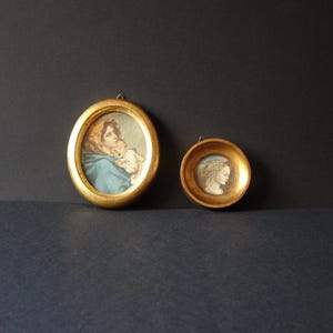 Op de afbeelding: Twee antieke miniatuurportretten in sierlijke gouden lijsten. Eén ovale lijst toont een vrouw in een blauwe sjaal die een kind vasthoudt. De andere, een kleinere ronde lijst, toont een vrouw in profiel. Beide worden tentoongesteld tegen een donkere achtergrond.