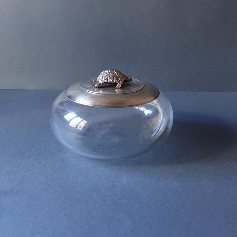 May include: A clear glass jar with a silver lid featuring a turtle figurine. The jar is round and has a smooth surface.