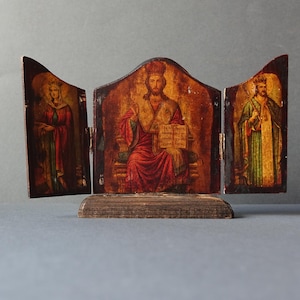 May include: A wooden triptych icon with three panels. The center panel depicts a figure in red robes holding a book. The left panel depicts a figure in a blue robe. The right panel depicts a figure in a green robe. The icon is on a wooden base.