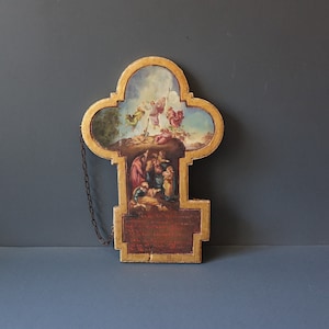 Op de afbeelding: Een houten, goud geverfde, kruisvormige plaquette met een religieuze scène erop geschilderd. De scène toont een groep mensen die rond een figuur op de grond zijn verzameld. Boven hen stijgen drie figuren op naar de hemel. De plaquette heeft een ketting die aan de bovenkant is bevestigd.