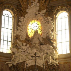 May include: An ornate, white religious altar with a stained glass window and a statue of a person in the centre. The altar is decorated with gold accents and candles.