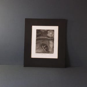 May include: A framed black and white etching print. The artwork depicts a boat on water under a moonlit sky. The print is matted in white and framed in a black frame. The artwork is displayed on a dark gray surface.