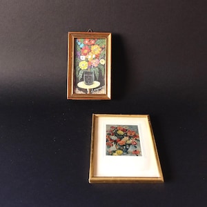 May include: Two framed floral paintings. The top painting features a vase of colorful flowers on a small table. The bottom painting shows a vase of flowers with a gold frame. Both paintings are displayed against a dark background.