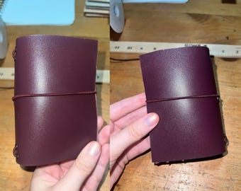 May include: A burgundy leather journal with a matching elastic closure. The journal is rectangular and has rounded corners. The elastic band wraps around the journal to keep it closed. The journal is shown on a wooden surface.